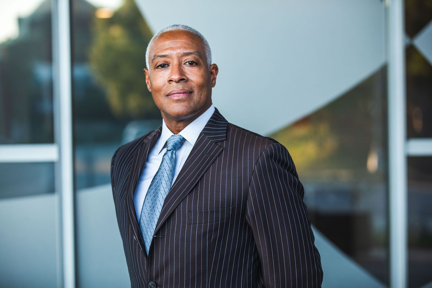 Professional executive in a pinstripe suit and blue tie poses for a corporate headshot in front of modern architecture.