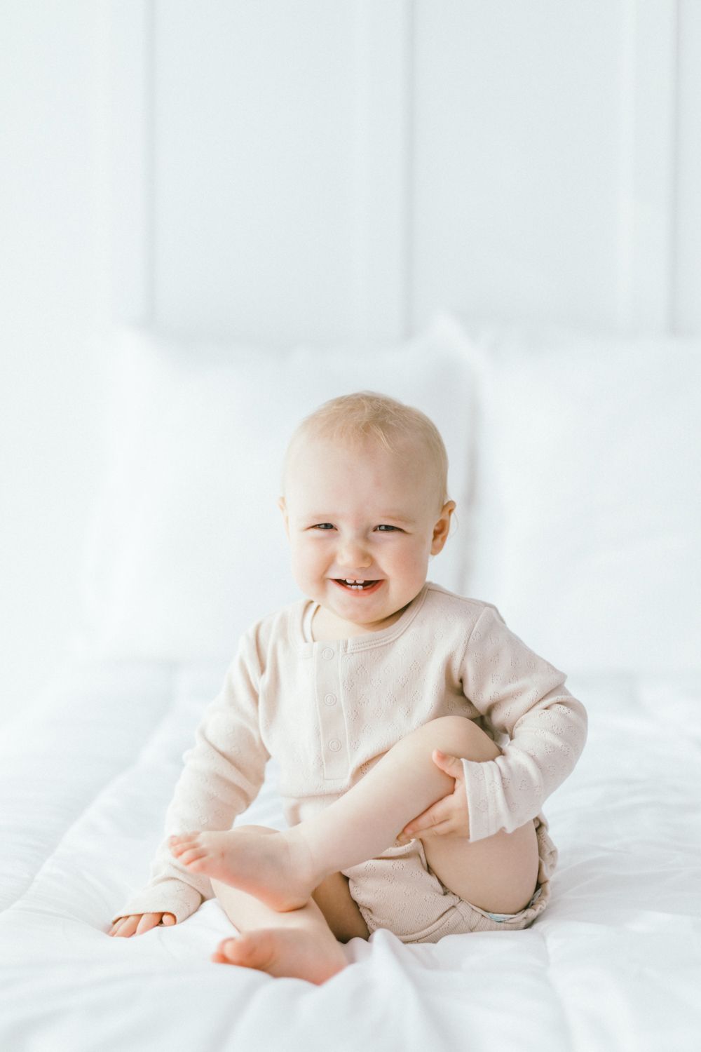 Joyful baby in beige outfit sitting on white bedding.