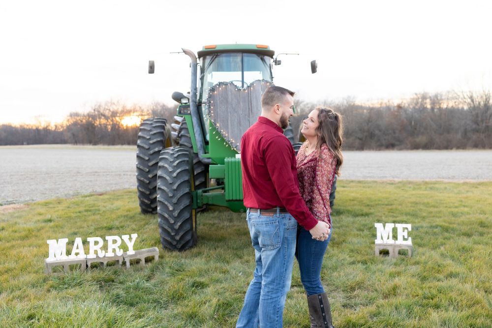 A couple stands in front of a large green tractor with 'MARRY ME' signs during a sunset proposal.