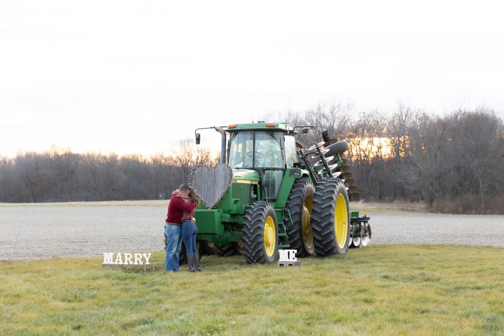 A green John Deere tractor in a field at sunset with 'MARRY ME' signs displayed in the grass.