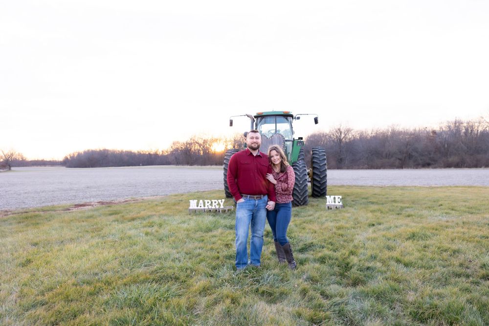 A couple poses for an engagement photo with a green tractor in a field at sunset.