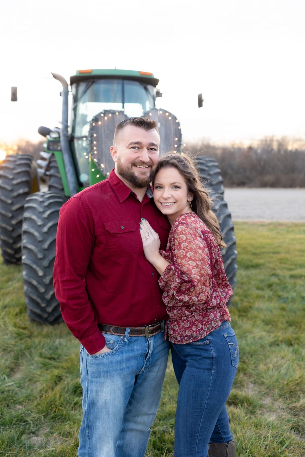 A happy couple poses together in front of a green tractor during their engagement shoot.
