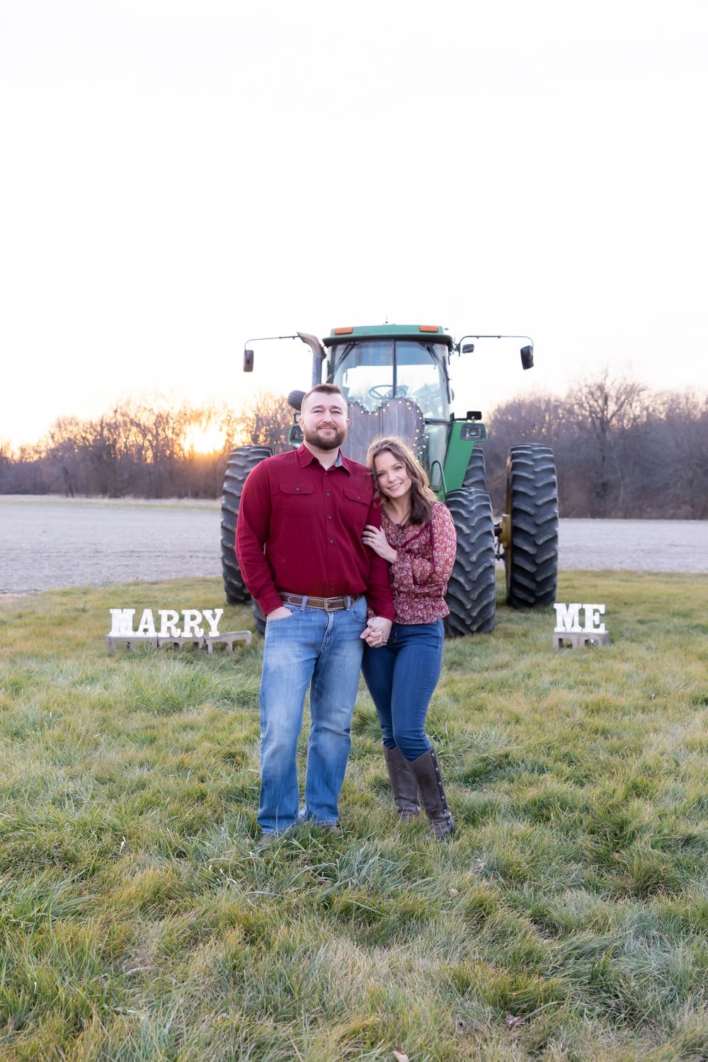 A couple stands together in front of a John Deere tractor with 'MARRY ME' signs visible.
