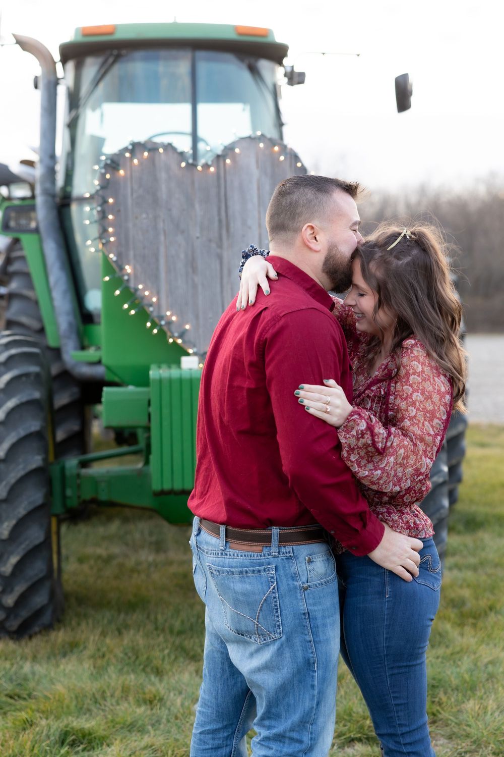 A couple embraces by a John Deere tractor decorated with fairy lights in a heart shape.