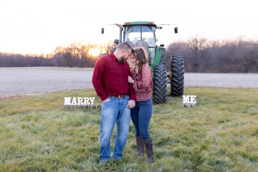 A couple shares a romantic moment in front of a tractor with 'MARRY ME' signs.