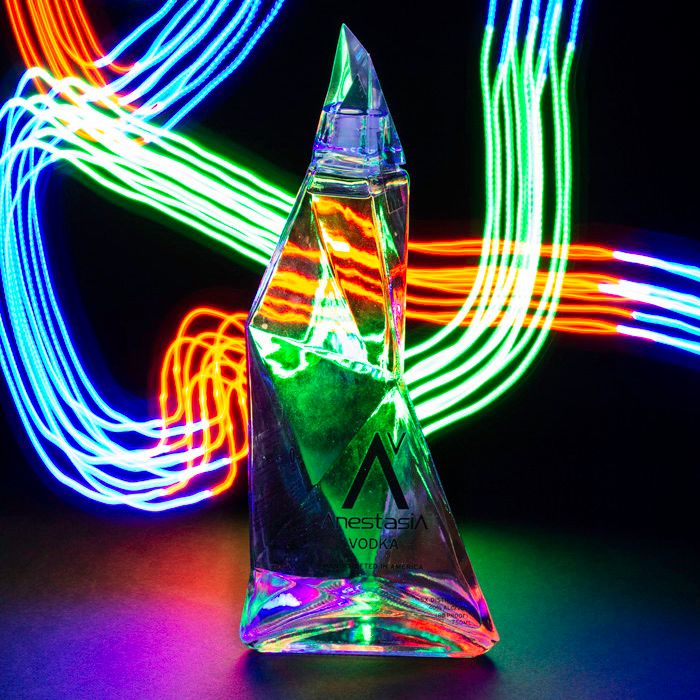 Colorful light painting surrounds a crystal pyramid bottle creating vibrant neon streaks against black background.