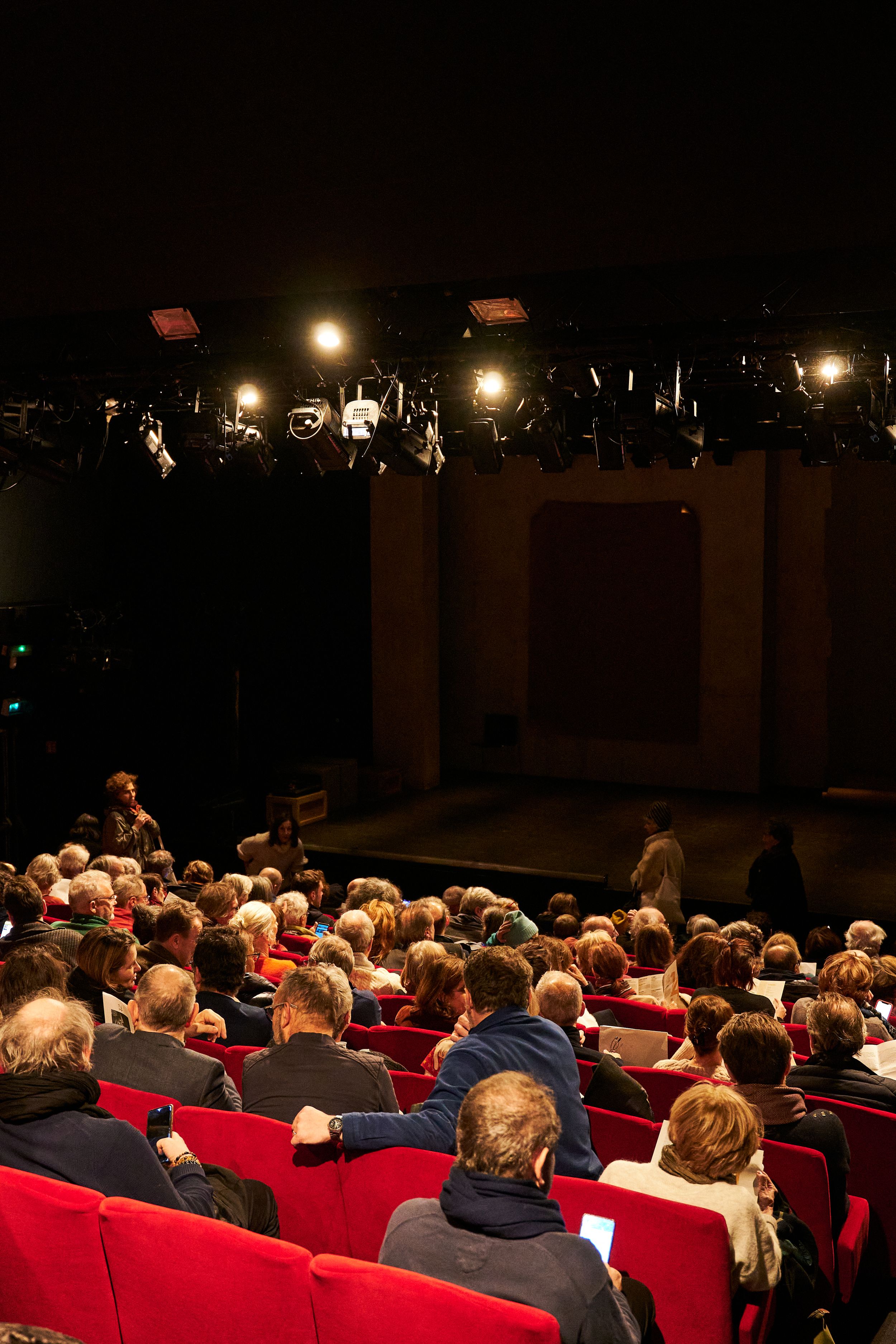 Theater audience seated in red chairs facing illuminated stage.