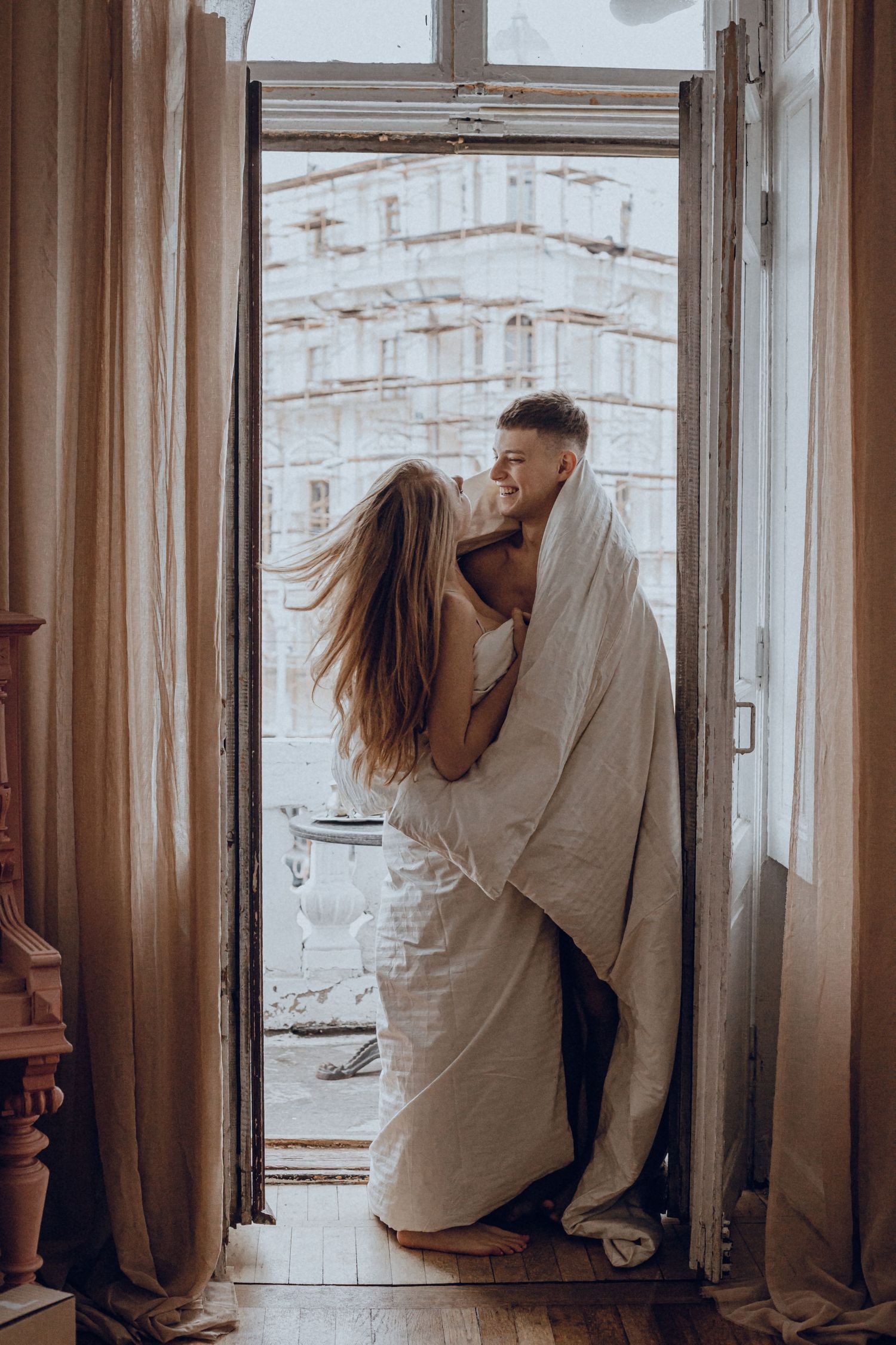 Couple wrapped in blanket sharing intimate moment by window in natural light.