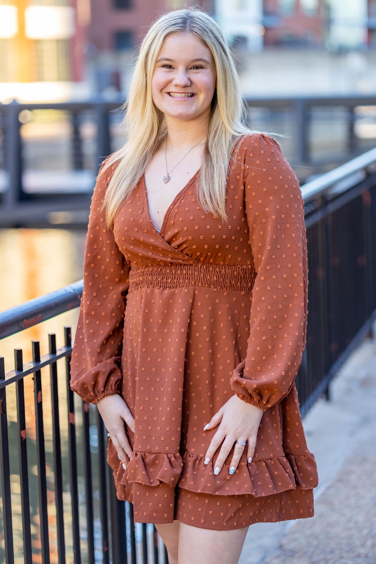 Person in rust-colored polka dot wrap dress with long sleeves standing by railing at sunset.