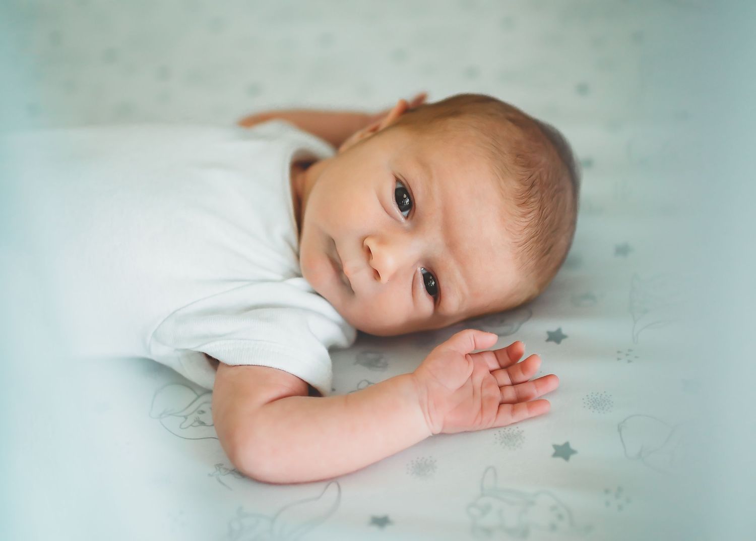 A baby lies on soft mint green bedding wearing a white outfit, looking directly at the camera.
