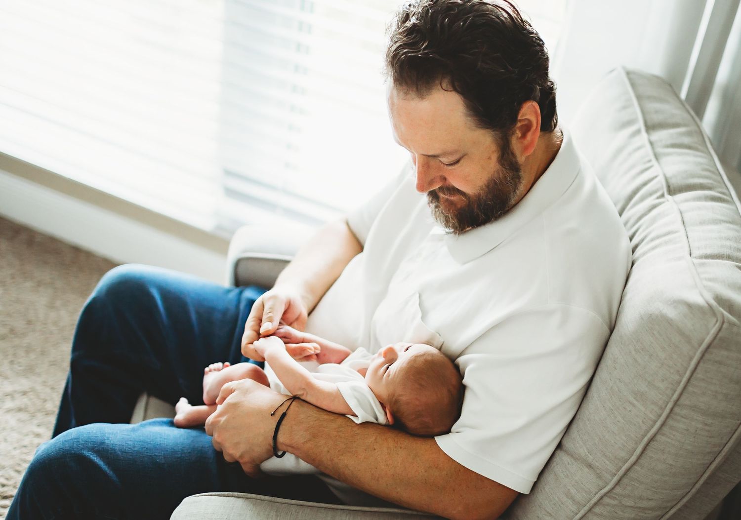 Bearded man in white shirt sits on couch while bottle feeding newborn baby in natural light setting.