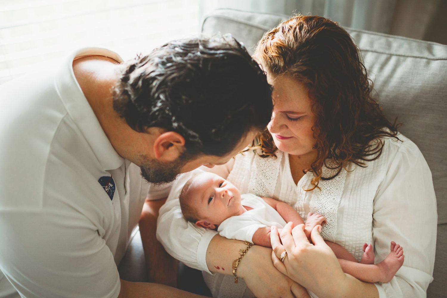 Two people in white clothing tenderly gazing at their newborn baby in a warm, intimate family moment.