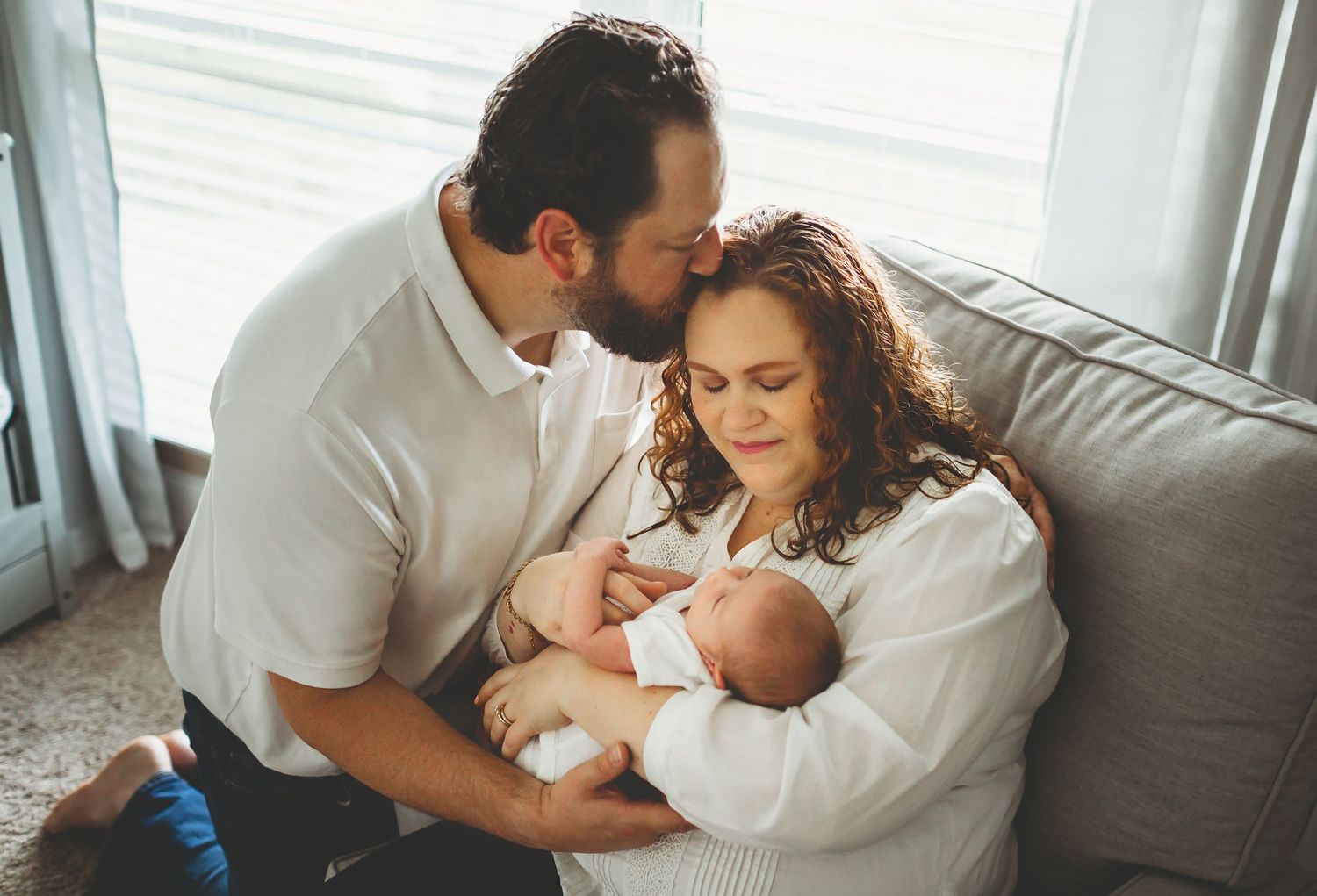 A tender moment shared between parents with their newborn baby while sitting together on a grey couch.
