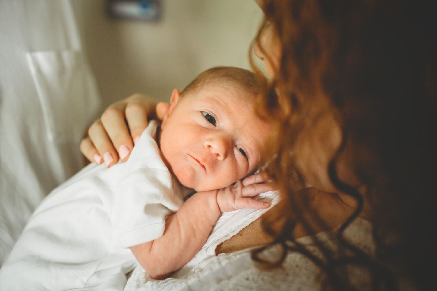 A baby in a white outfit rests comfortably while being held in soft light.