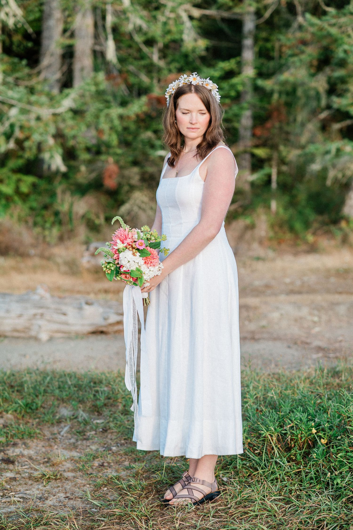 bride in white dress holding floral bouquet stands in grassy outdoor setting with trees in background.