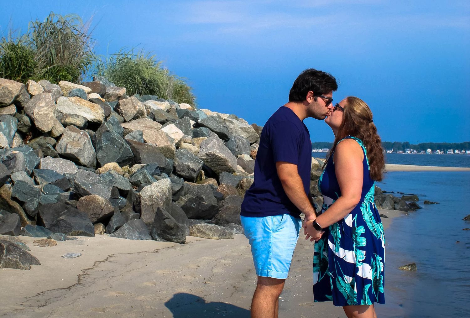 A romantic couple shares a tender moment on a rocky beach with blue ocean waters in the background.
