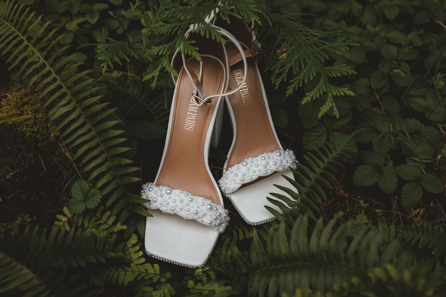 White bridal shoes nestled among green ferns and foliage.