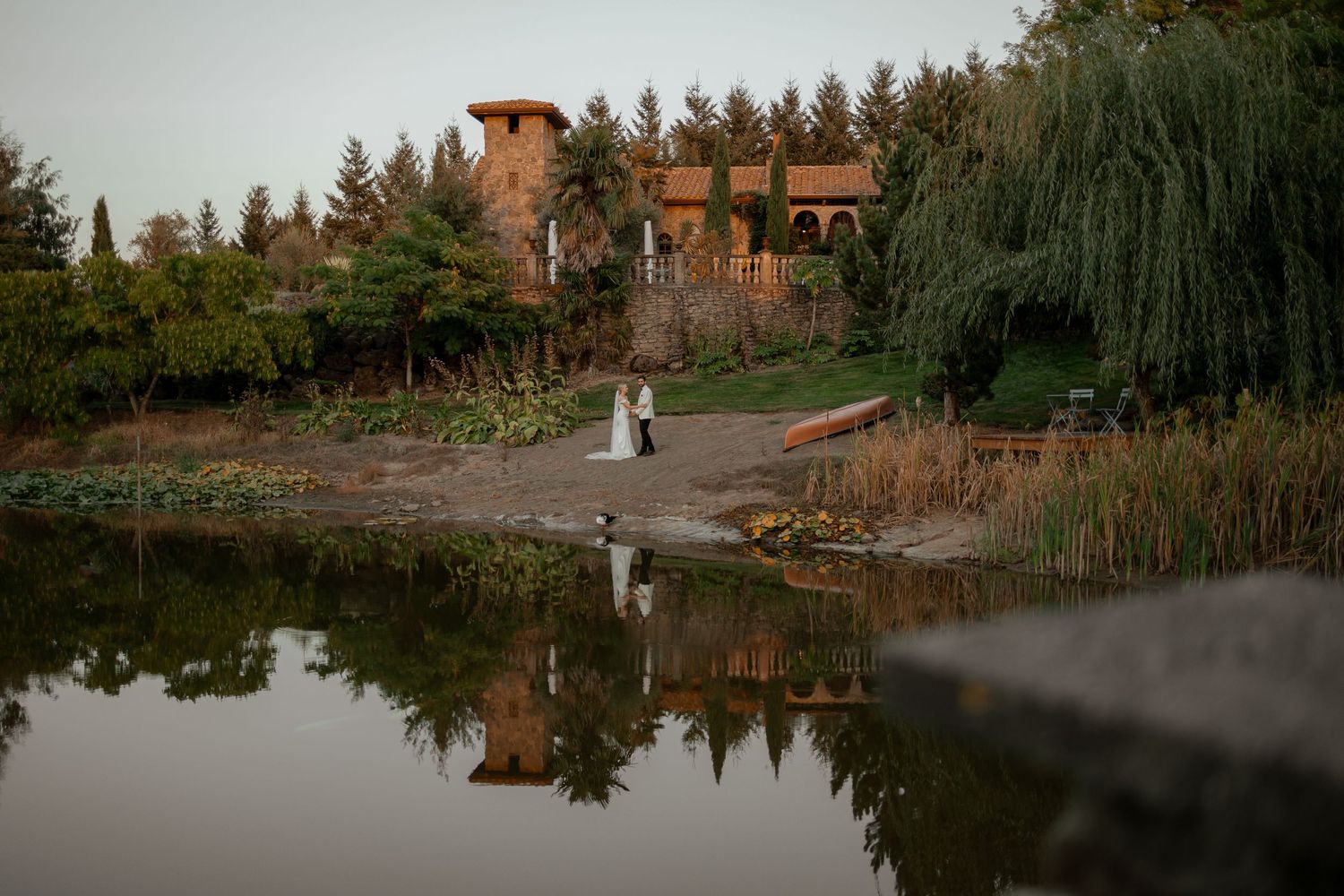 Couple walking near a reflective pond with rustic building and trees in background at sunset.