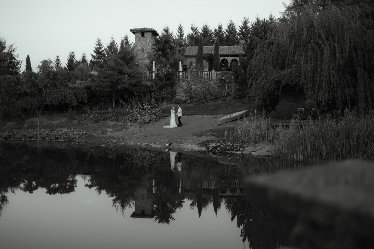 Black and white image of figures by a pond with building and willow trees reflected in still water.