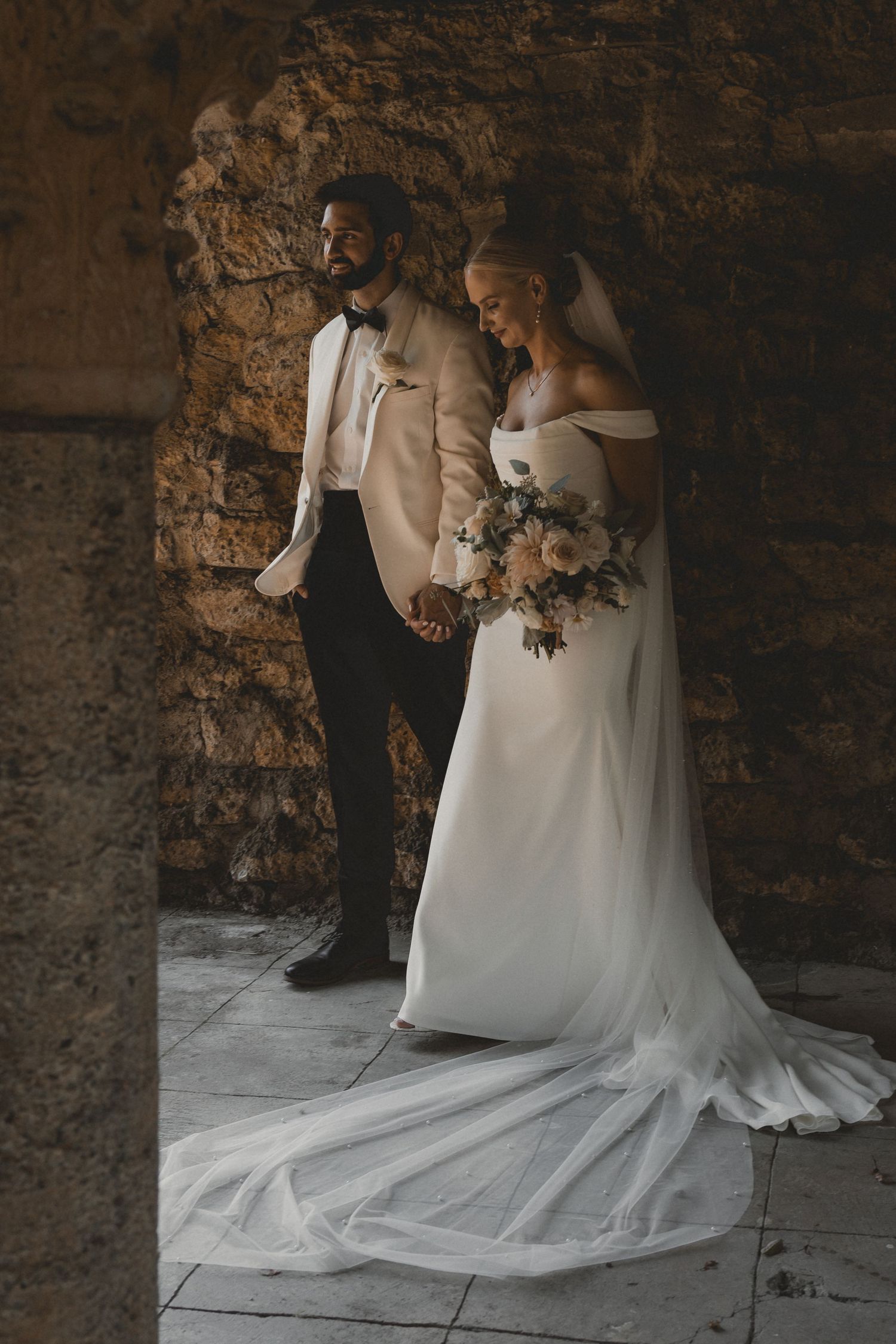 A romantic wedding photo showing a couple in formal attire against a rustic stone wall backdrop.