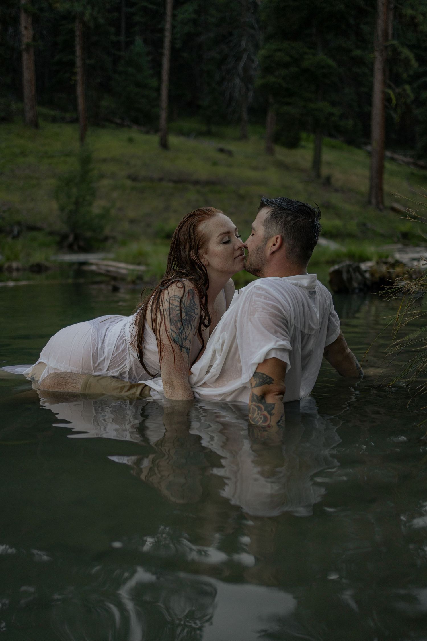 A romantic couple shares an intimate kiss while standing in a serene lake surrounded by forest.