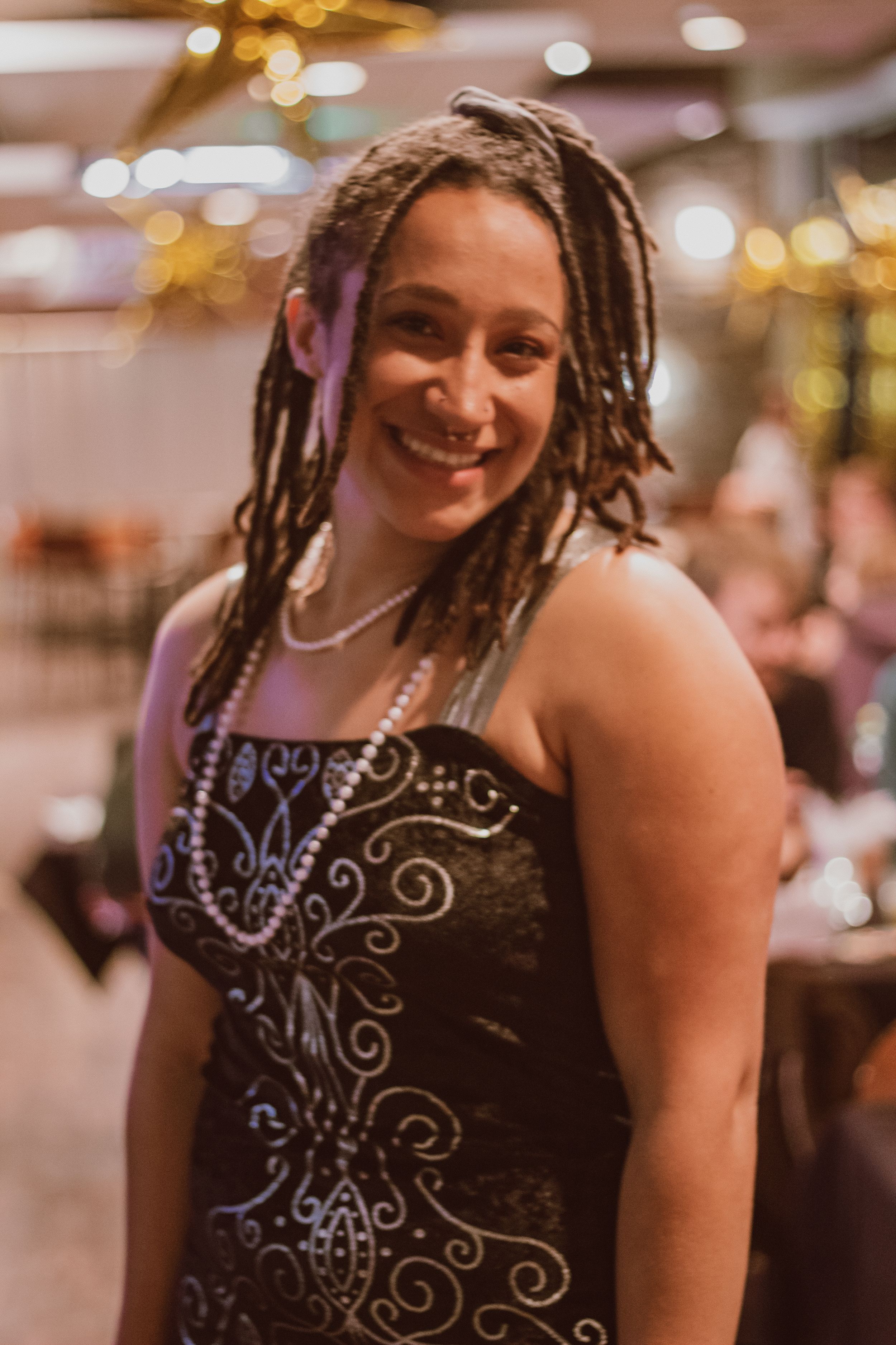 A person wearing a black dress with decorative swirl patterns and beaded necklaces at an indoor event.
