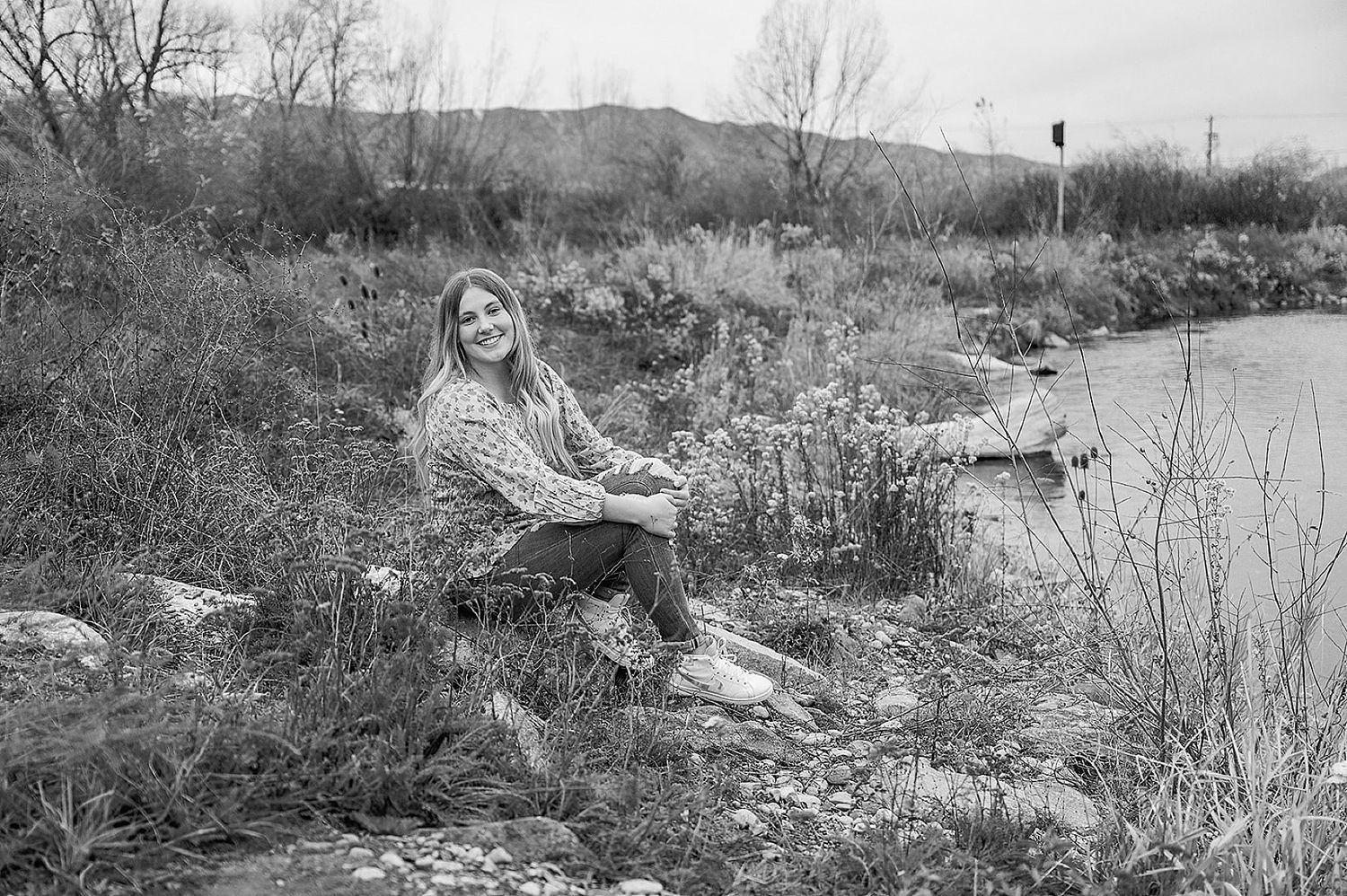 Black and white photograph of someone sitting on rocks by a riverbank surrounded by autumn vegetation.