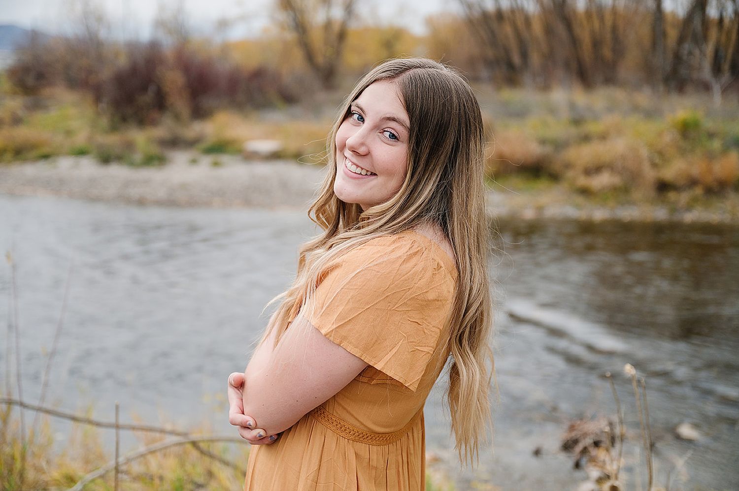 Casual outdoor portrait near a river with autumn foliage and mountains in background.