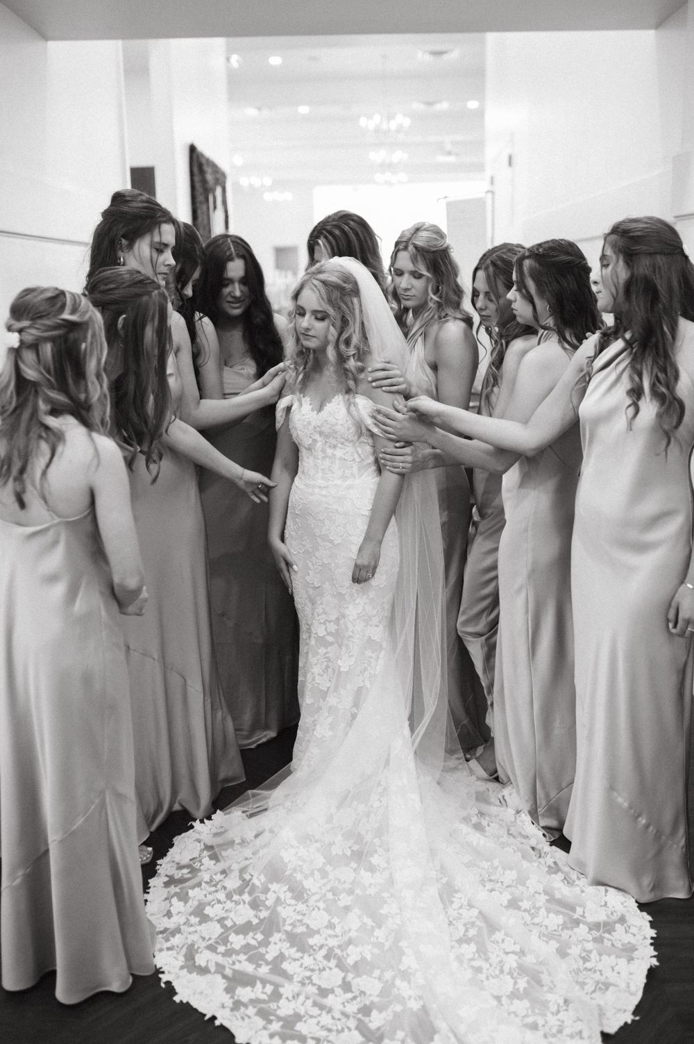 A group gathered around in sleek silk dresses helps adjust a stunning lace wedding gown with a dramatic train.