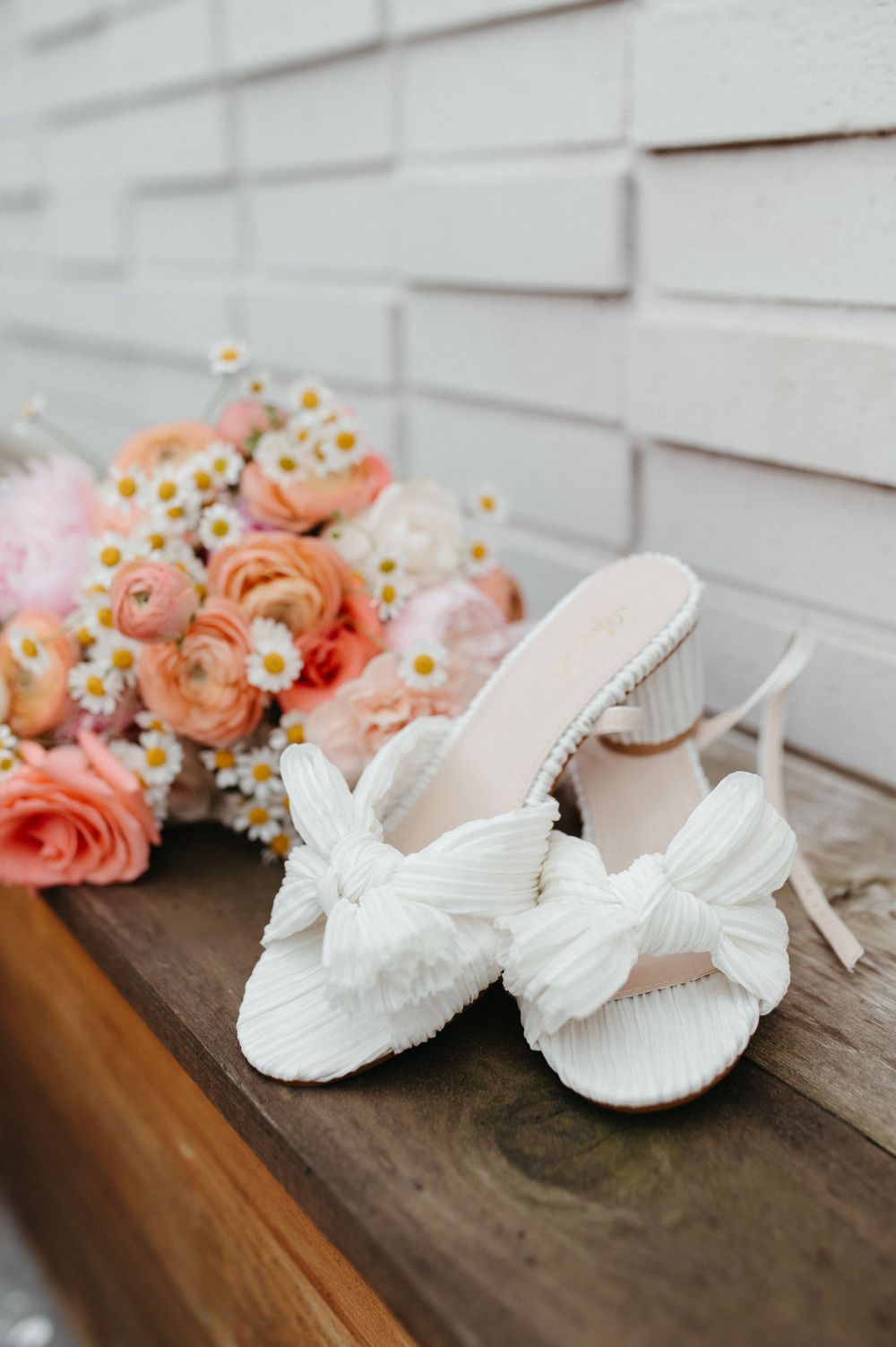 White bridal bow heels displayed with coral roses and daisies on wooden surface.