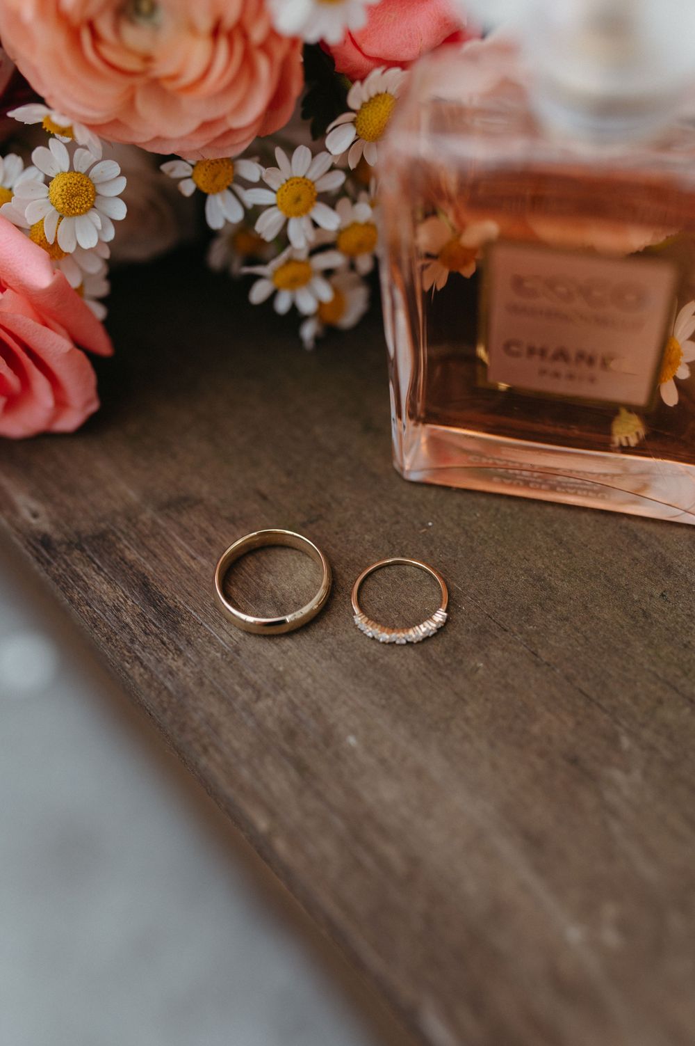 Wedding rings beside pink roses, daisies and perfume bottle on wooden surface.