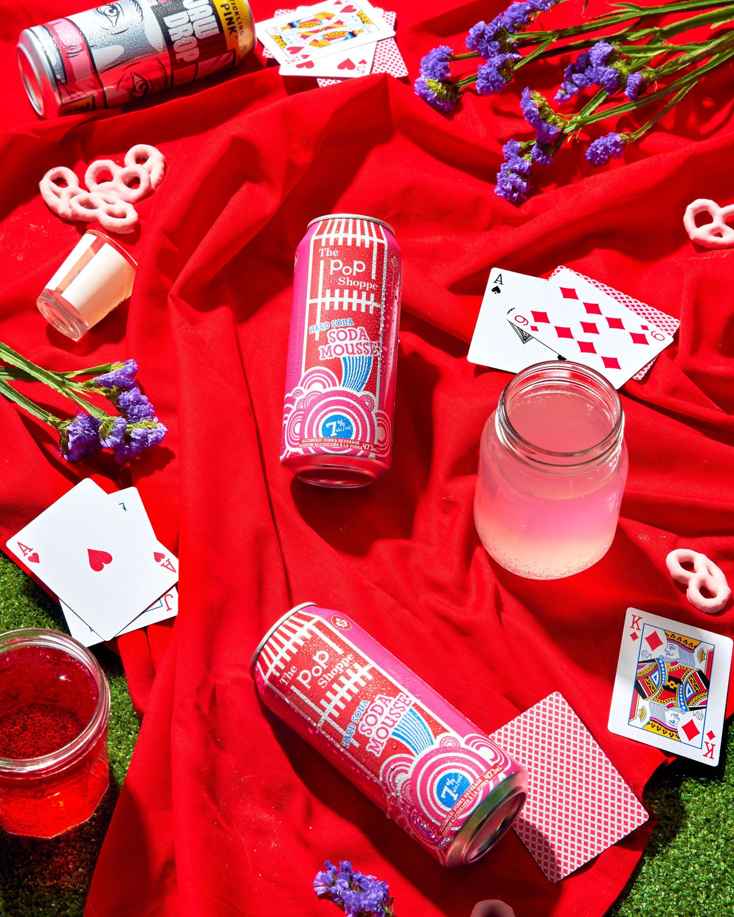 Playing cards and pink drinks scattered on red fabric with grass visible at the edges create a festive scene.