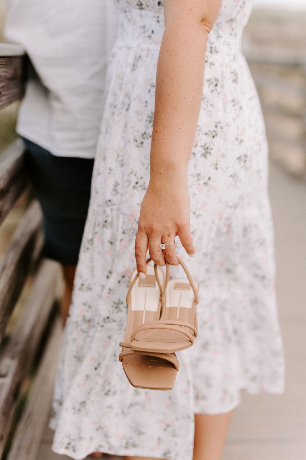 Hand holding small tan purse wearing floral dress against white background.