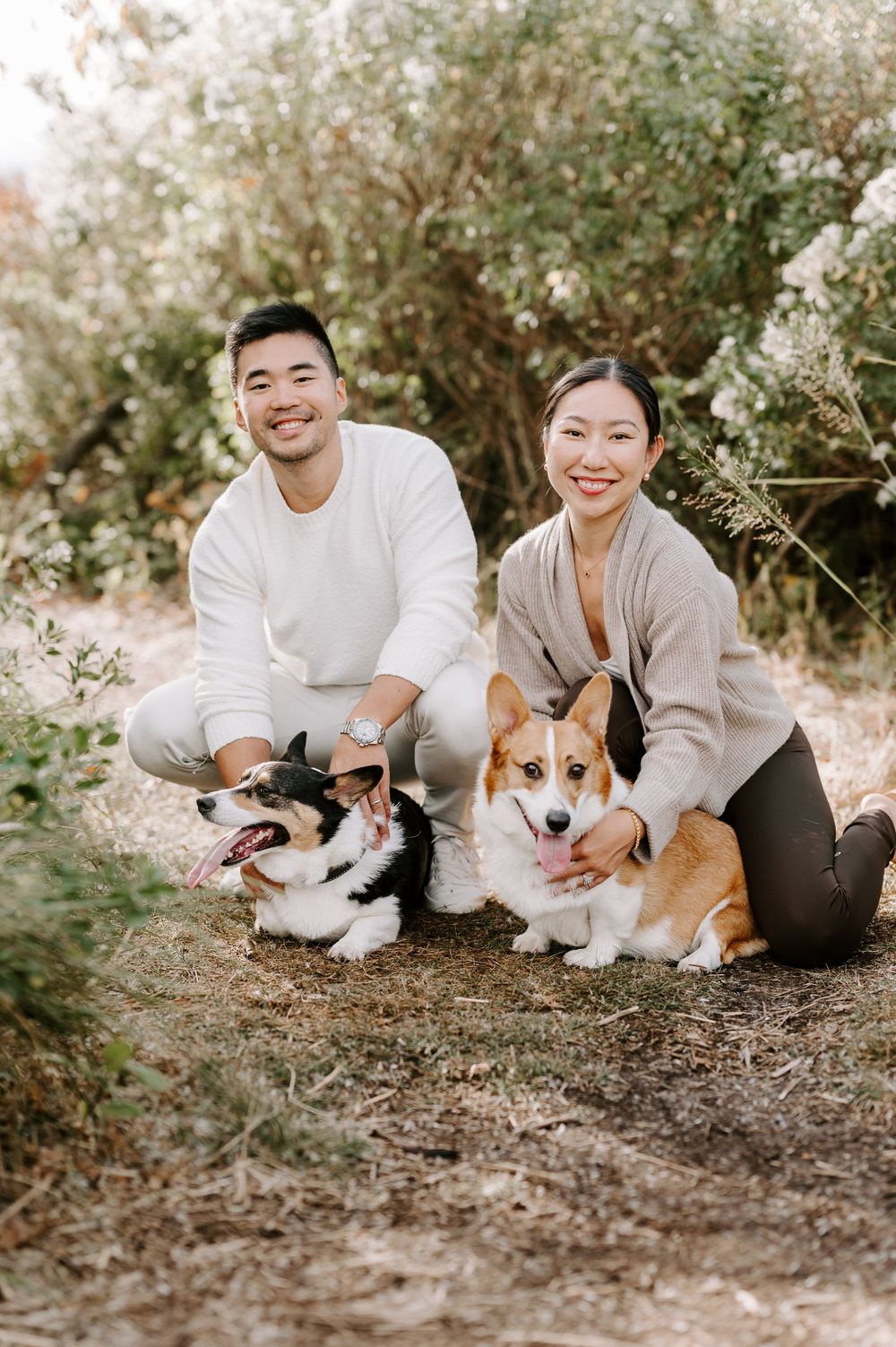 Couple in neutral sweaters poses with their two Corgi dogs on nature trail.