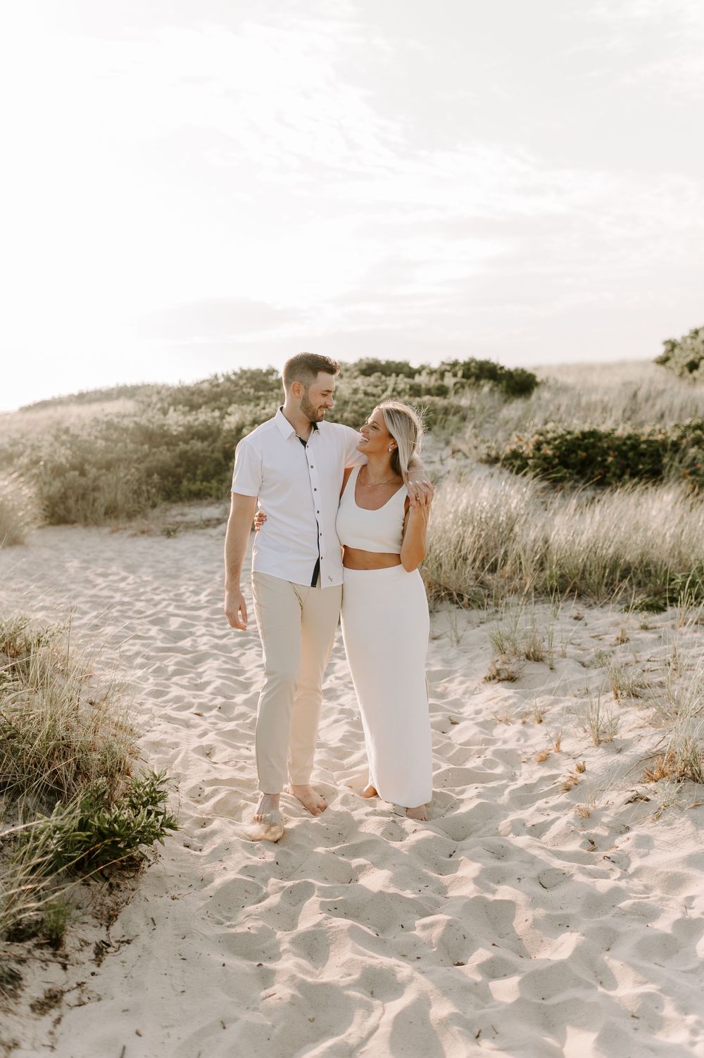 A couple in white outfits walks hand in hand along a sandy beach path at sunset.
