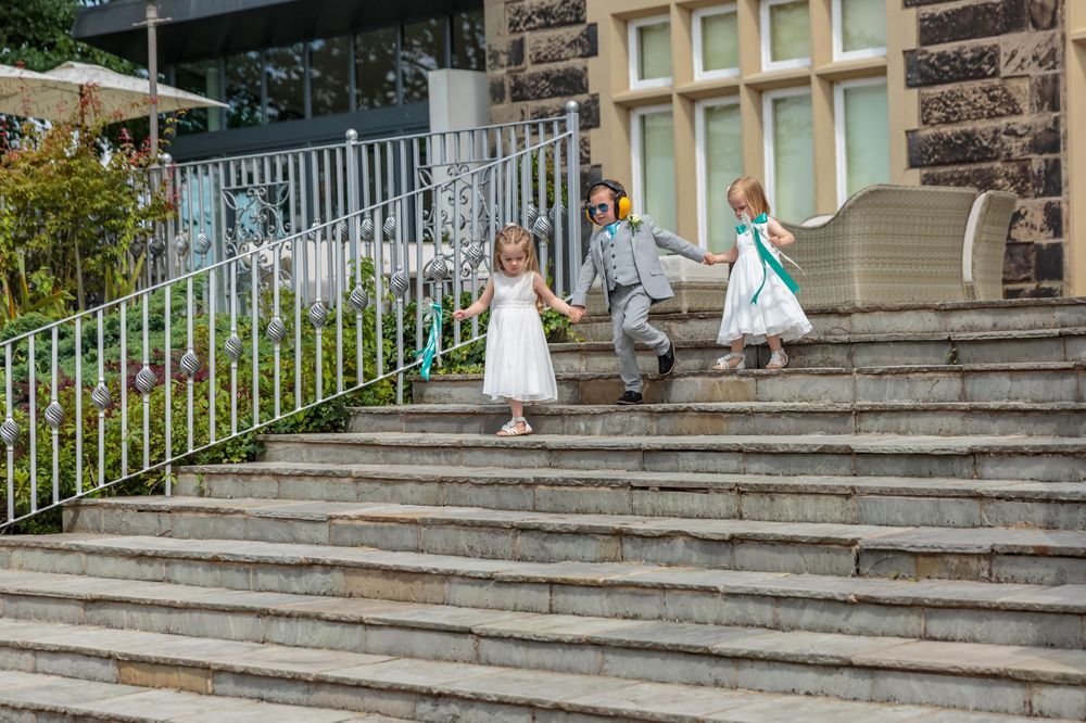 young page boy wearing ear defenders walking down the steps outside west tower holding hands with the flower girls