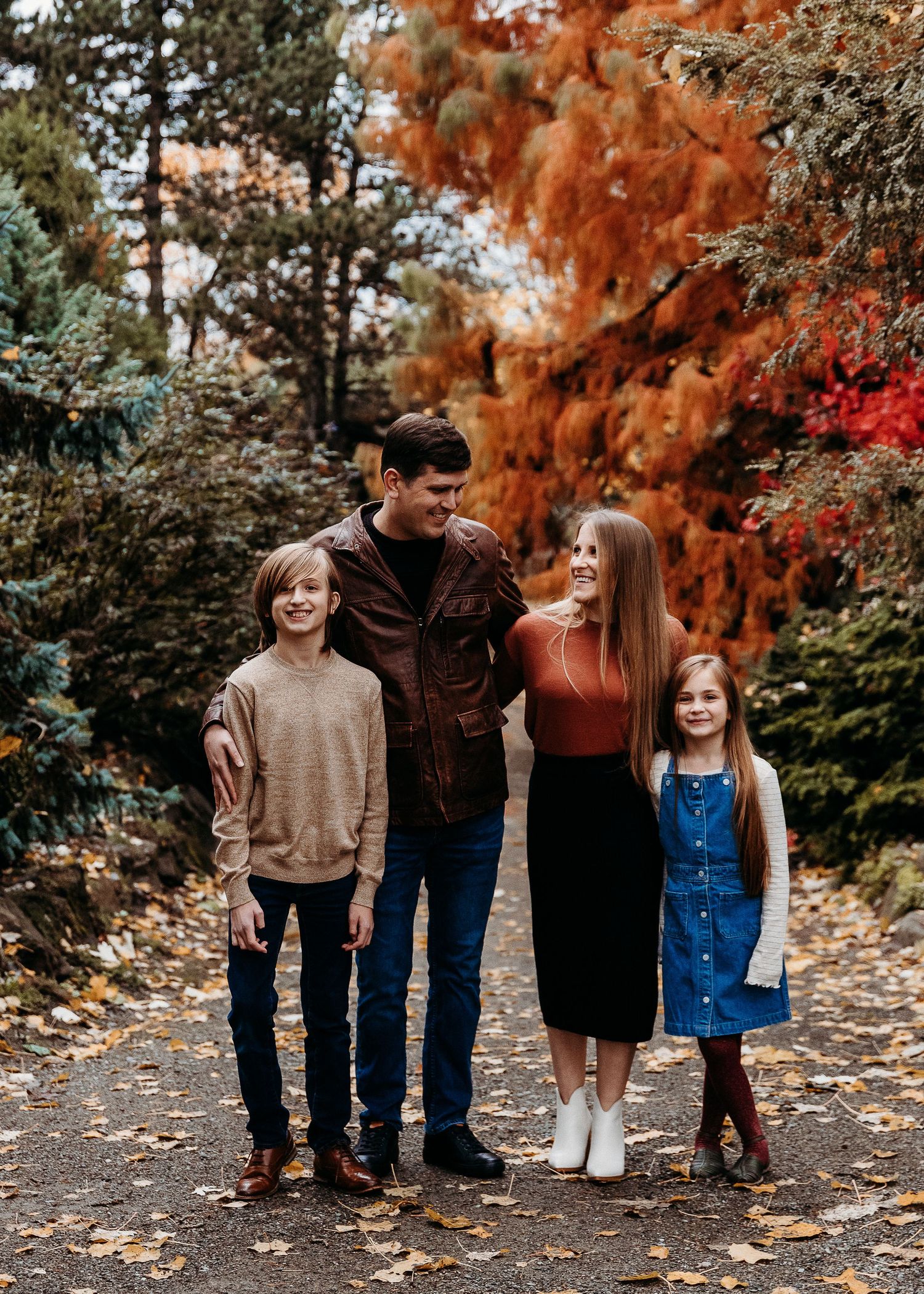 A family in coordinated autumn outfits poses on a leaf-covered path surrounded by colorful trees.