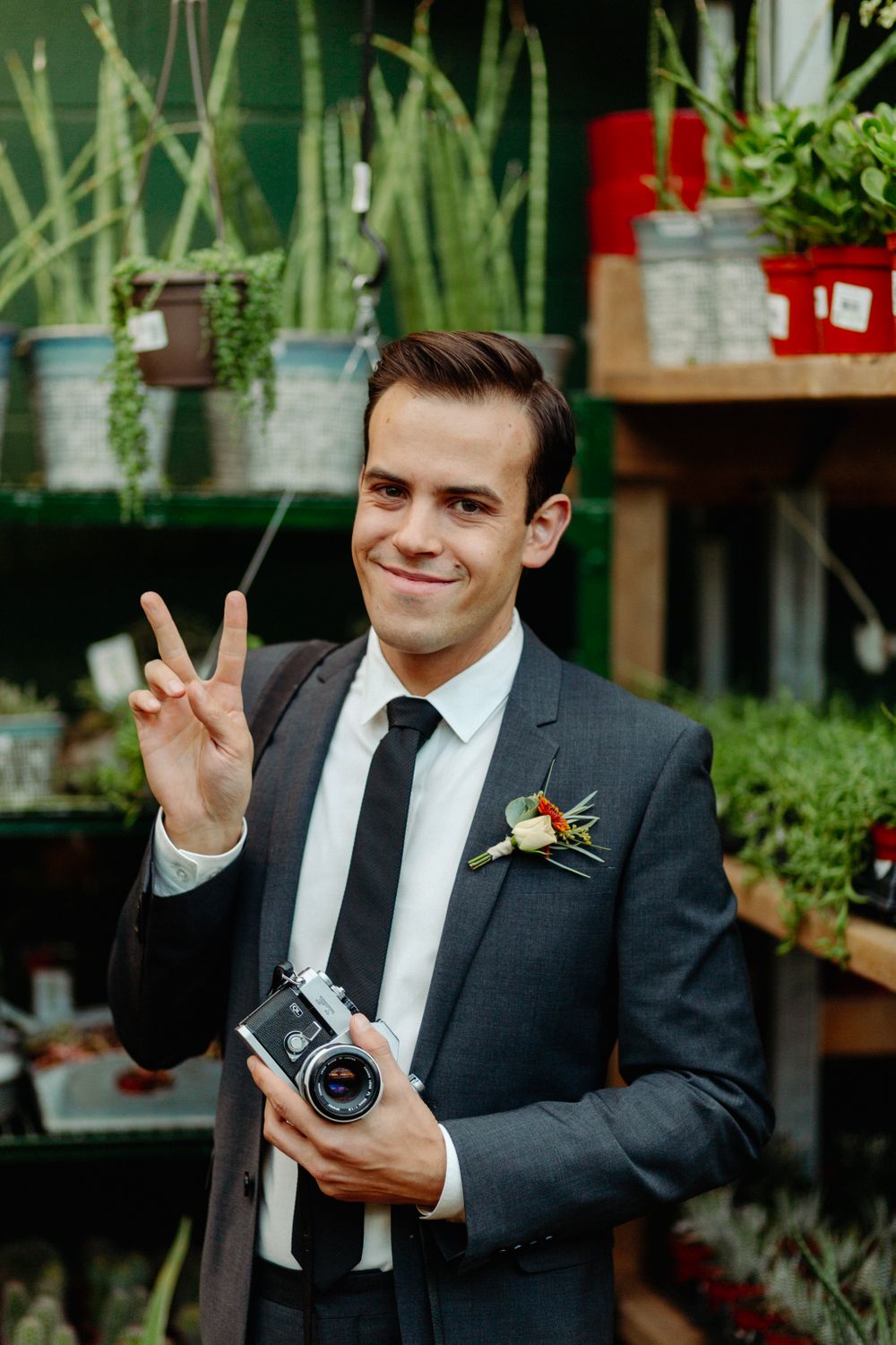 A suited figure with vintage camera making peace sign in front of plant display.