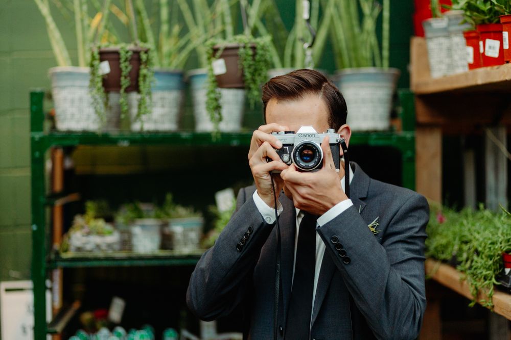A photographer in formal attire taking pictures among garden plants and greenery.