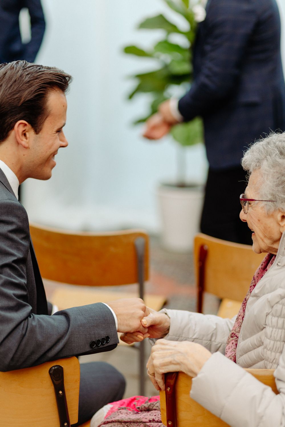 A person in a suit speaking warmly with an elderly woman wearing glasses in an indoor setting.