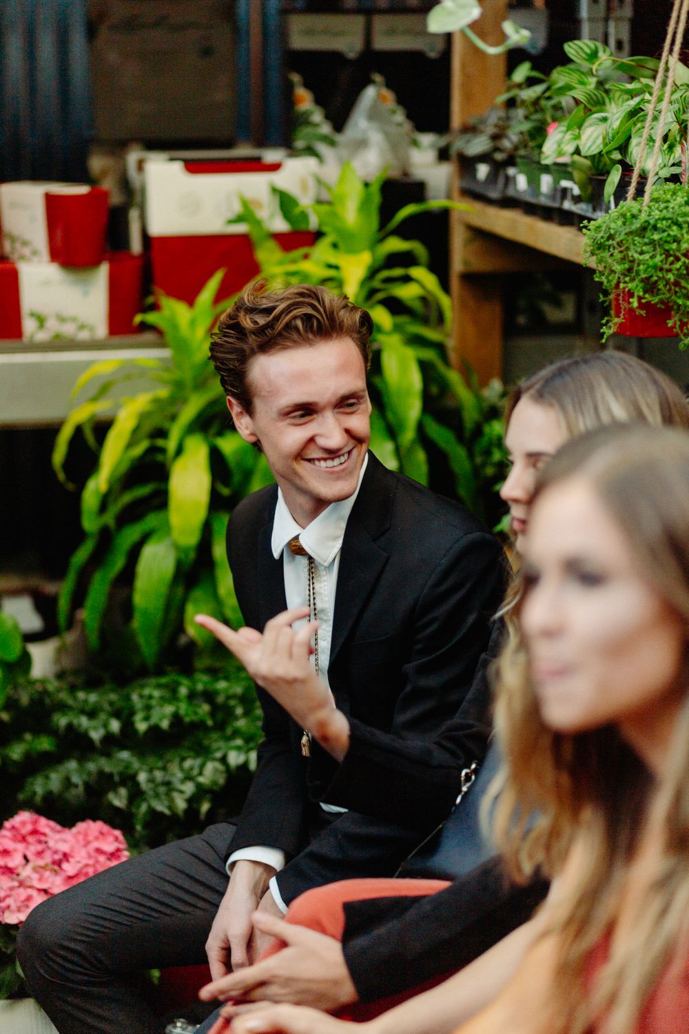 A person in formal attire smiling brightly among indoor plants at a social gathering.