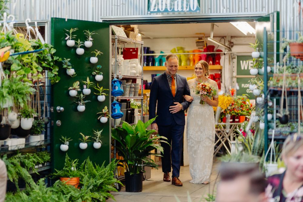 Couple enters a brightly lit flower shop decorated with plants and colorful flowers.