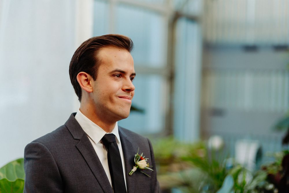 A groom in a dark suit smiles during an indoor garden wedding ceremony.