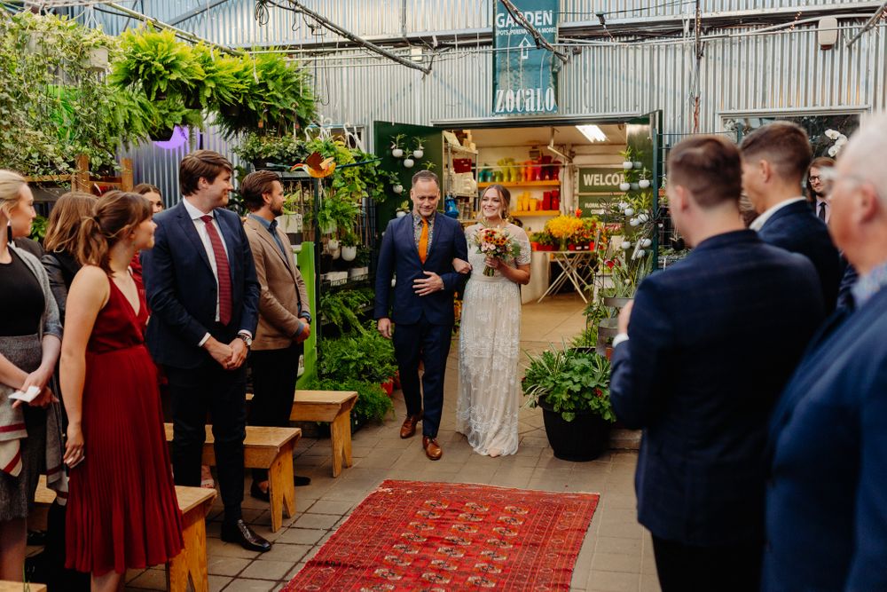 Indoor greenhouse wedding ceremony with guests gathered on a red carpet aisle surrounded by plants and greenery.
