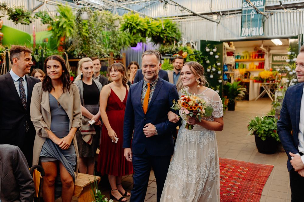 Wedding guests gather in a greenhouse-style venue filled with plants and flowers.
