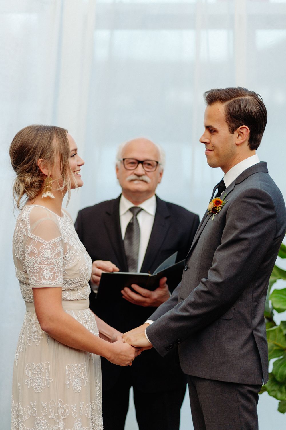 Indoor greenhouse wedding ceremony with white backdrop and potted tropical plants.