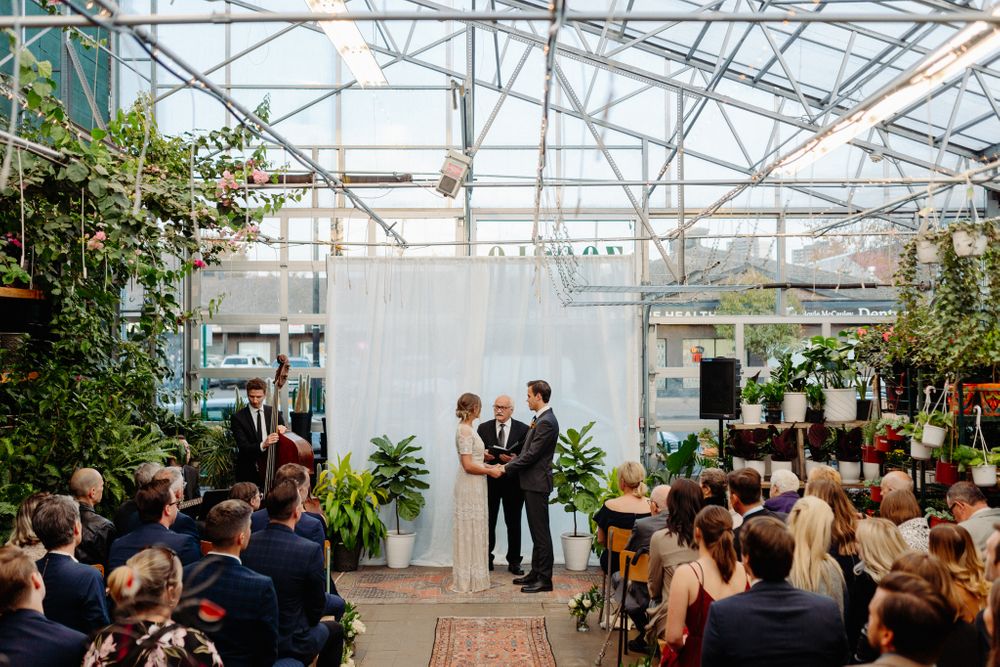 Intimate wedding ceremony in greenhouse with white backdrop and tropical plant decor.