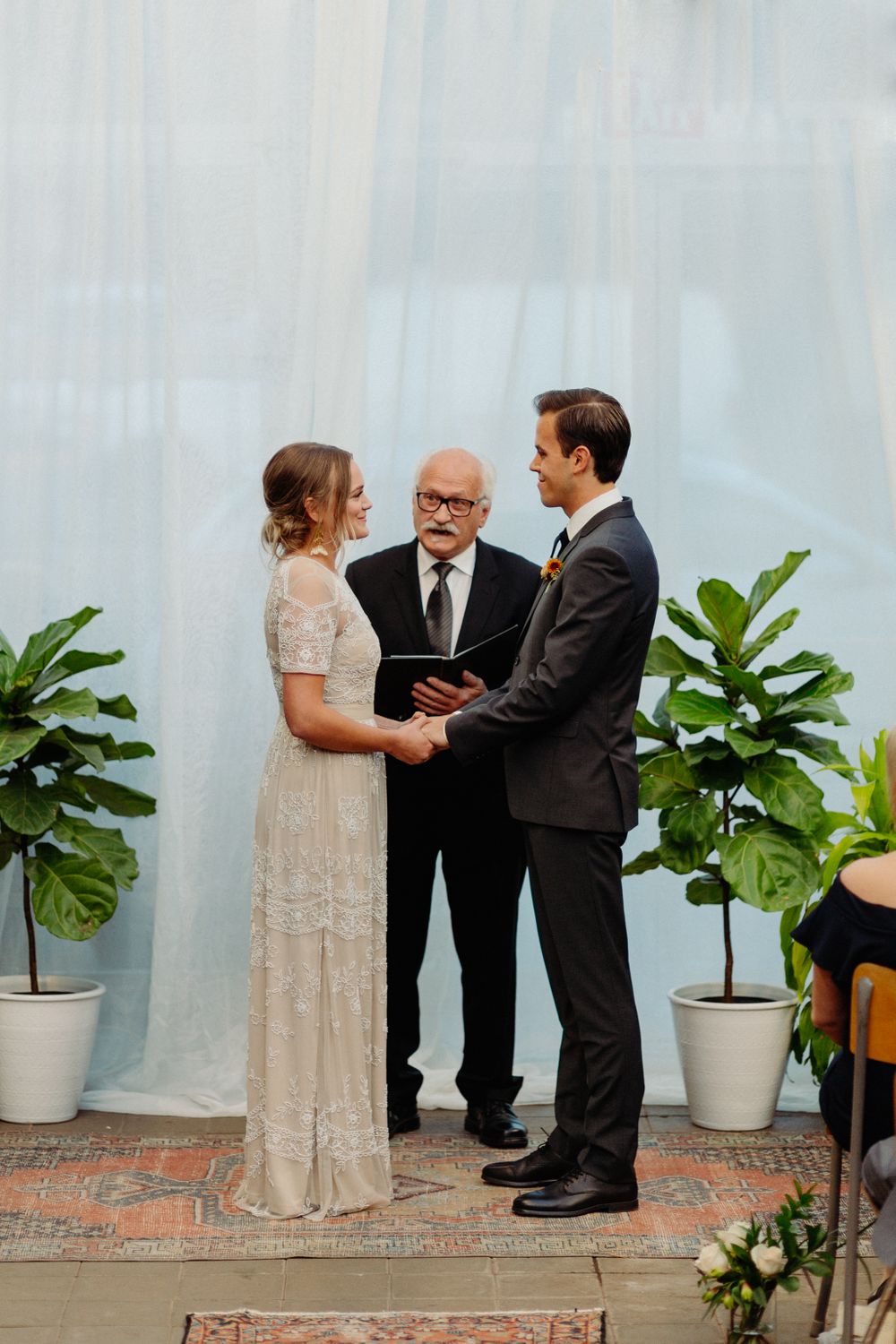 A wedding ceremony in progress inside a greenhouse with potted plants and white curtains.
