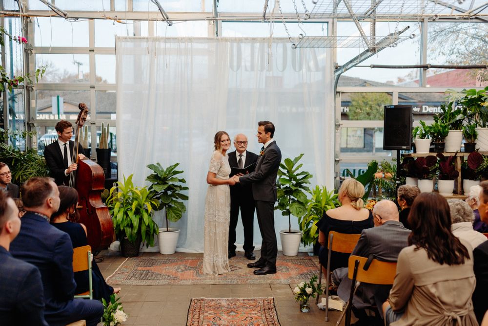 An intimate wedding ceremony takes place in a greenhouse-style venue with white curtains and potted plants.
