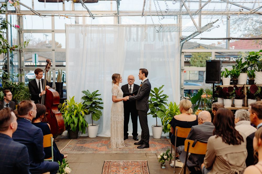 A wedding ceremony unfolds in a bright greenhouse space with guests seated on wooden chairs.